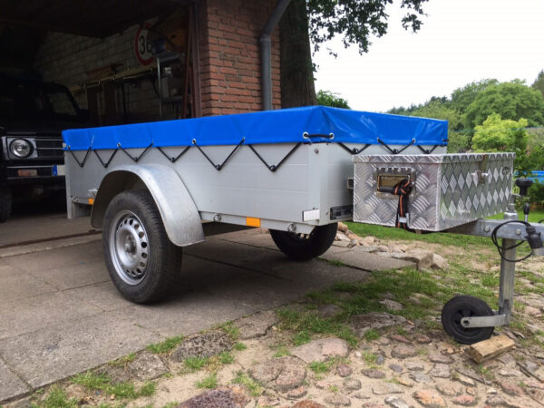 Ein kleiner Mehrzweckanhänger mit einer blauen Flachplane-Plane und einem Werkzeugkasten aus Metall an der Vorderseite steht in der Nähe einer gemauerten Garage auf einer gepflasterten Auffahrt, umgeben von Grün.