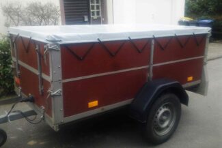 Ein kleiner brauner Anhänger mit weißer Flachplane und schwarzen Einachsrädern, geparkt in einem Wohngebiet. Mit schwarzen Schnüren gesichert und mit Seitenreflektoren ausgestattet.