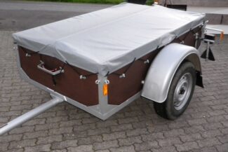 Ein kleiner kastenförmiger Anhänger auf grauen Steinen hat ein mit schwarzen Gummiseilen sicher befestigtes Flachflugzeug. Er verfügt über eine silberne Plane, eine braune Karosserie und einen roten Griff über der Einzelachse.