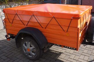 Ein kleiner orangefarbener Nutzanhänger verfügt über ein Flachflugzeug mit schwarzen Kabeln, einen schwarzen Kotflügel und ein Reserverad an der Seite. Er steht auf einem Bürgersteig mit Grünflächen und einer Mauer.