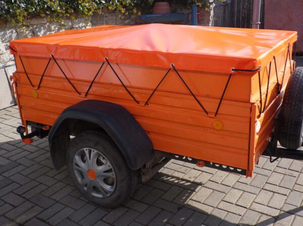 Ein kleiner orangefarbener Nutzanhänger verfügt über ein Flachflugzeug mit schwarzen Kabeln, einen schwarzen Kotflügel und ein Reserverad an der Seite. Er steht auf einem Bürgersteig mit Grünflächen und einer Mauer.