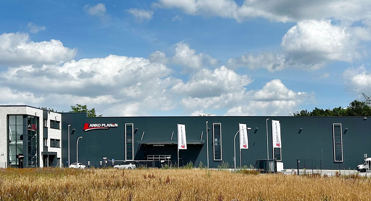 Ein modernes Industriegebäude mit weißen und dunkelgrauen Abschnitten, Fahnen am Eingang und einer Wiese davor unter einem blauen Himmel mit vereinzelten Wolken.