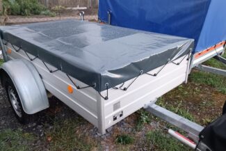 Ein mit einer grauen Flachplane bedeckter Mehrzweckanhänger steht auf dem Gras neben einem anderen mit einer blauen Abdeckung. Er verfügt über einen Metallrahmen und ein einzelnes sichtbares Rad. Im Hintergrund ist ein Zaun zu sehen.