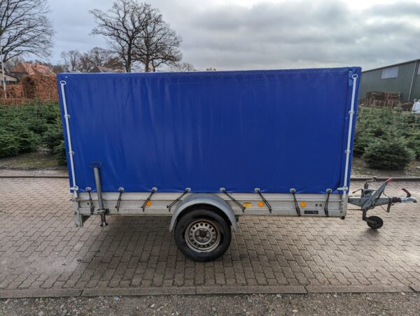 Ein kleiner einrädriger Pritschenanhänger mit einer leuchtend blauen Hochplane-Abdeckung steht auf einem gepflasterten Platz im Freien, umgeben von Bäumen und einem bewölkten Himmel.