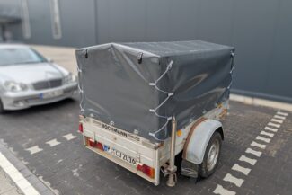 Ein kleiner überdachter Anhänger mit einem Hochplane mit Spitzdach und einer grauen Plane ist auf einem Parkplatz neben einem silbernen Auto geparkt, das das Bockmann-Logo auf der Rückseite zeigt.