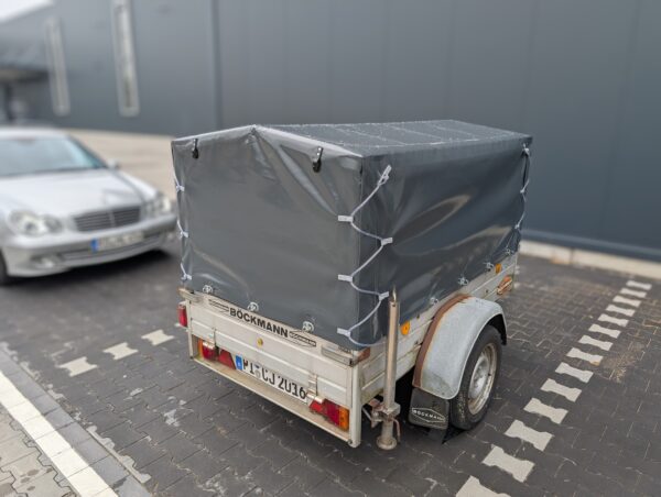 Ein kleiner überdachter Anhänger mit einem Hochplane mit Spitzdach und einer grauen Plane ist auf einem Parkplatz neben einem silbernen Auto geparkt, das das Bockmann-Logo auf der Rückseite zeigt.