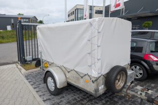 Ein kleiner Anhänger mit einer weißen Hochplane-Plane ist neben einem roten Auto und einem Metallzaun geparkt, im Hintergrund sind Gebäude und Fahnen zu sehen.