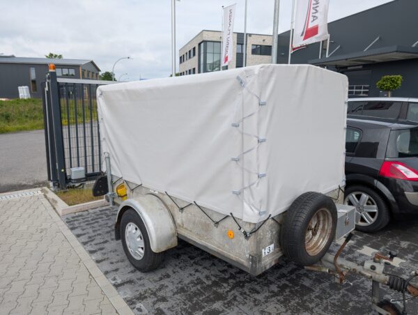 Ein kleiner Anhänger mit einer weißen Hochplane-Plane ist neben einem roten Auto und einem Metallzaun geparkt, im Hintergrund sind Gebäude und Fahnen zu sehen.