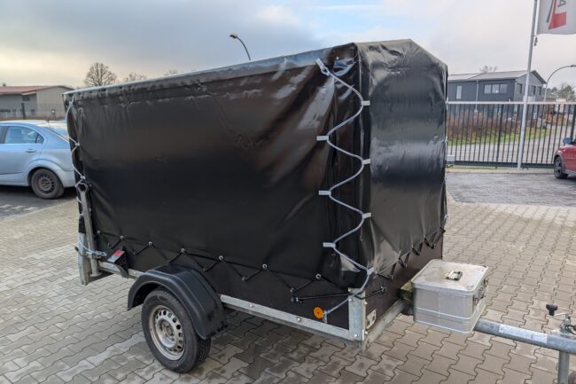Ein kleiner Anhänger mit Hochplane mit Spitzdach und schwarzer Plane ist auf einem gepflasterten Platz in der Nähe eines silbernen Autos und von Gebäuden abgestellt.