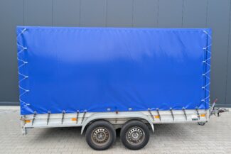 Ein silberner zweiachsiger Anhänger mit einem leuchtend blauen Hochplane-Logo steht auf hellen Pflastersteinen neben einer grauen Mauer.