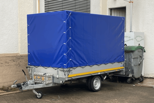 Ein Anhänger mit einem blauen Hochplane ist neben einem Metallcontainer vor einem Gebäude mit beigen und weißen Wänden geparkt.