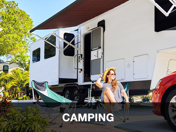 Eine Frau mit Sonnenbrille und Sonnenhut sitzt in einem Campingstuhl neben einem großen weißen Wohnmobil mit Markise. In der Nähe parkt ein rotes Auto, das teilweise von einem Anhängerplane bedeckt ist. Bäume und Pflanzen schaffen eine ruhige Kulisse. „CAMPING.