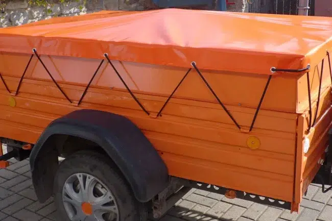 Flachplane-orange Orangefarbener Anhänger mit Plane, gesichert durch Seile. Er steht auf einer befestigten Fläche. Der Anhänger hat einen schwarzen Radkasten und ein Reserverad mit einer Radkappe mit orangefarbener Mitte.