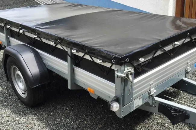 Flachplane-schwarz Auf dem Kies steht ein Nutzanhänger mit stabilem Metallrahmen und Anhängerplane-Abdeckung. Er hat eine einzelne Achse und Räder, die Seitenwände sind aus gewellten Metallplatten gefertigt.