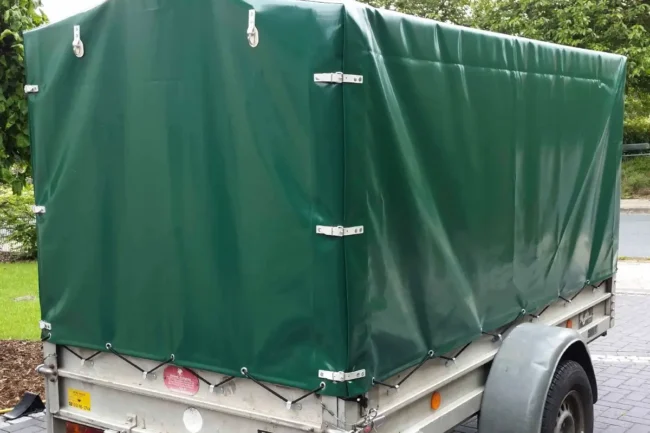 Hochplane-gruen Ein kleiner Nutzanhänger mit einer grünen Anhängerplane steht auf einer gepflasterten Fläche. Er hat eine einzelne Achse, sichtbare Räder und Metallbeschläge, die die Plane sichern. Bäume und Gras zieren den Hintergrund.