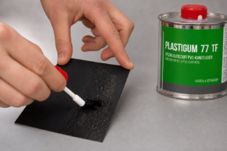 Eine Person streicht Kleber auf schwarzes Material; eine 250ml-Dose PVC-Planenkleber - Plastigum77 steht in der Nähe auf einer grauen Fläche.