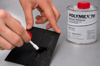 Mit einem Pinsel wird transparenter Klebstoff auf schwarzes Material aufgetragen. Daneben steht eine 250-ml-Pinseldose mit PVC-Kleber – Polymex70 – und rotem Deckel.