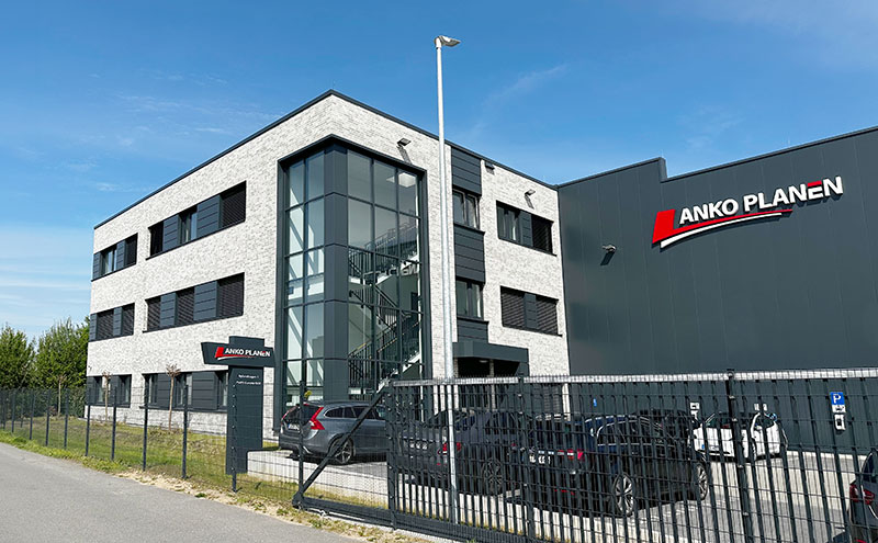 Ein modernes, graues Geschäftsgebäude mit ANKO PLANEN-Beschilderung, großen Fenstern, geparkten Autos und einem eingezäunten Gelände an einem sonnigen Tag.