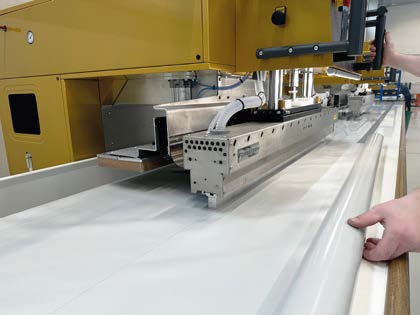 Eine Nahaufnahme einer Industriemaschine in einer Fabrik, die ein großes Blatt weißen Materials verarbeitet, wobei die Hände einer Person das Blatt führen.