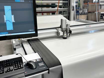 Eine computergesteuerte Schneidemaschine schneidet weißes Material zu, während ein Monitor das Schnittmuster anzeigt; eine Tastatur und eine Maus befinden sich auf einem Schreibtisch in der Nähe.