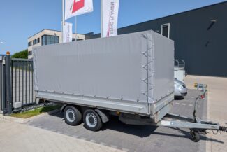 Ein grauer, überdachter Anhänger mit zwei Rädern ist auf einem gepflasterten Grundstück neben einem schwarzen Gebäude und einem silbernen Auto geparkt, in der Nähe befinden sich Fahnen und ein Zaun.